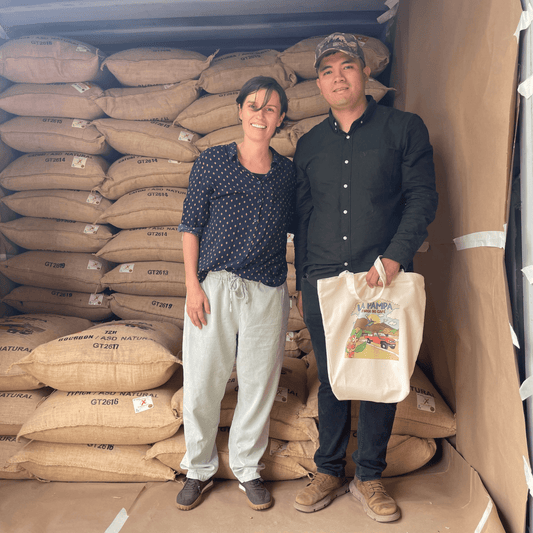 Mann und Frau vor Kaffeesäcken mit Tüte in der Hand. Es handelt sich um die Farmer*innen