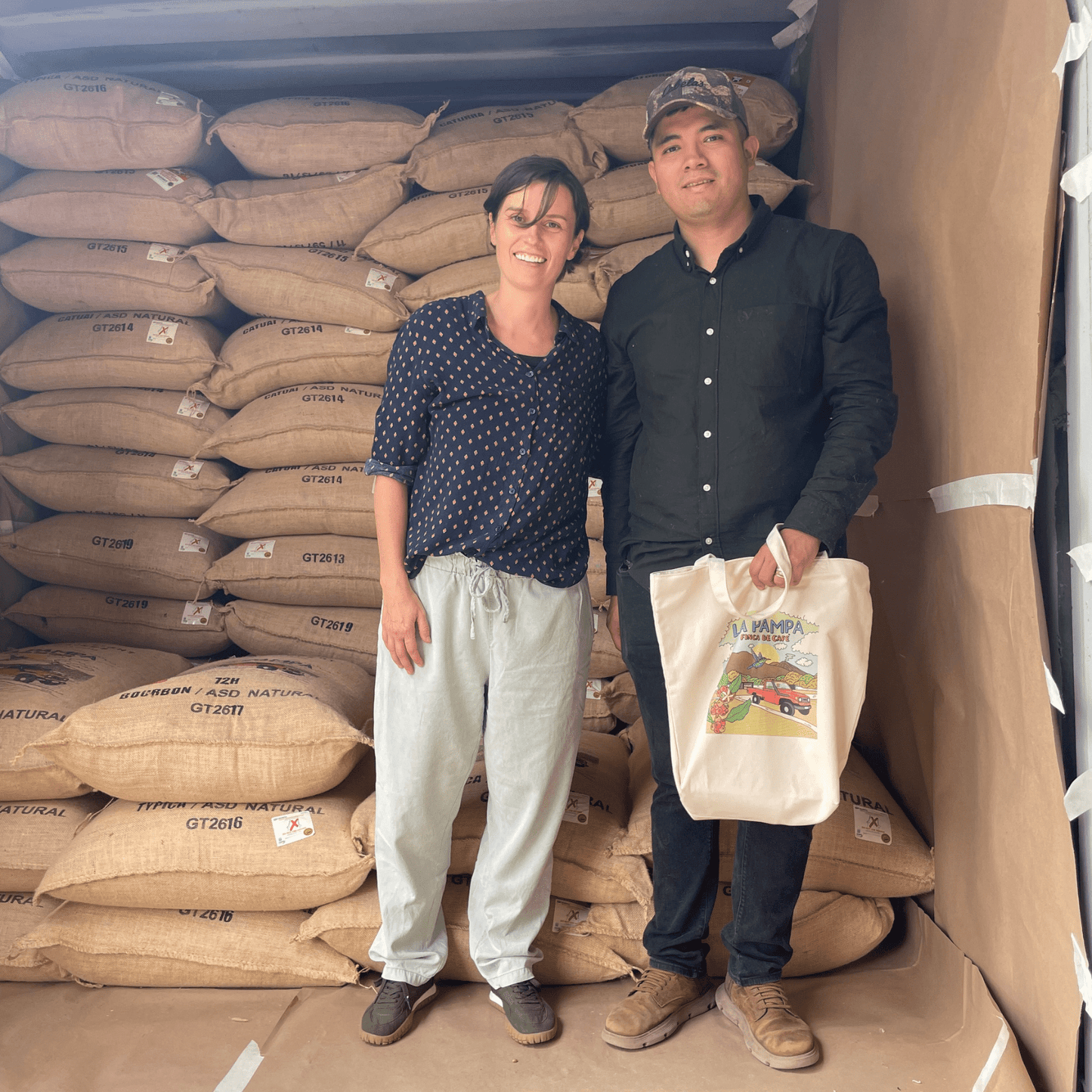 Mann und Frau vor Kaffeesäcken mit Tüte in der Hand. Es handelt sich um die Farmer*innen