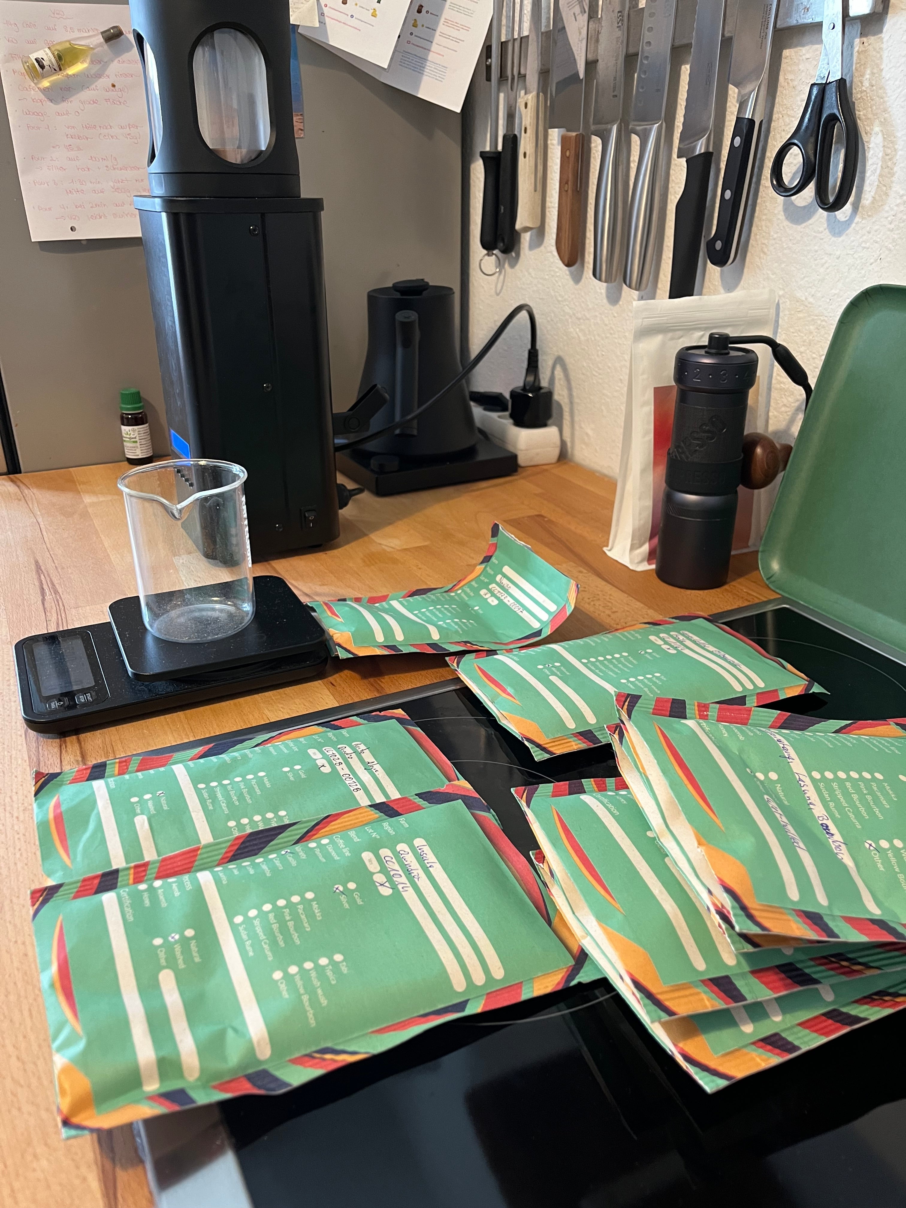 Verschiedene kleine Rohkaffeepackungen, auf Küchenarbeitsplatte. Kleiner Sampleröster im Hintergrund. Wasserkocher und Handmühle rechts im Bild.
