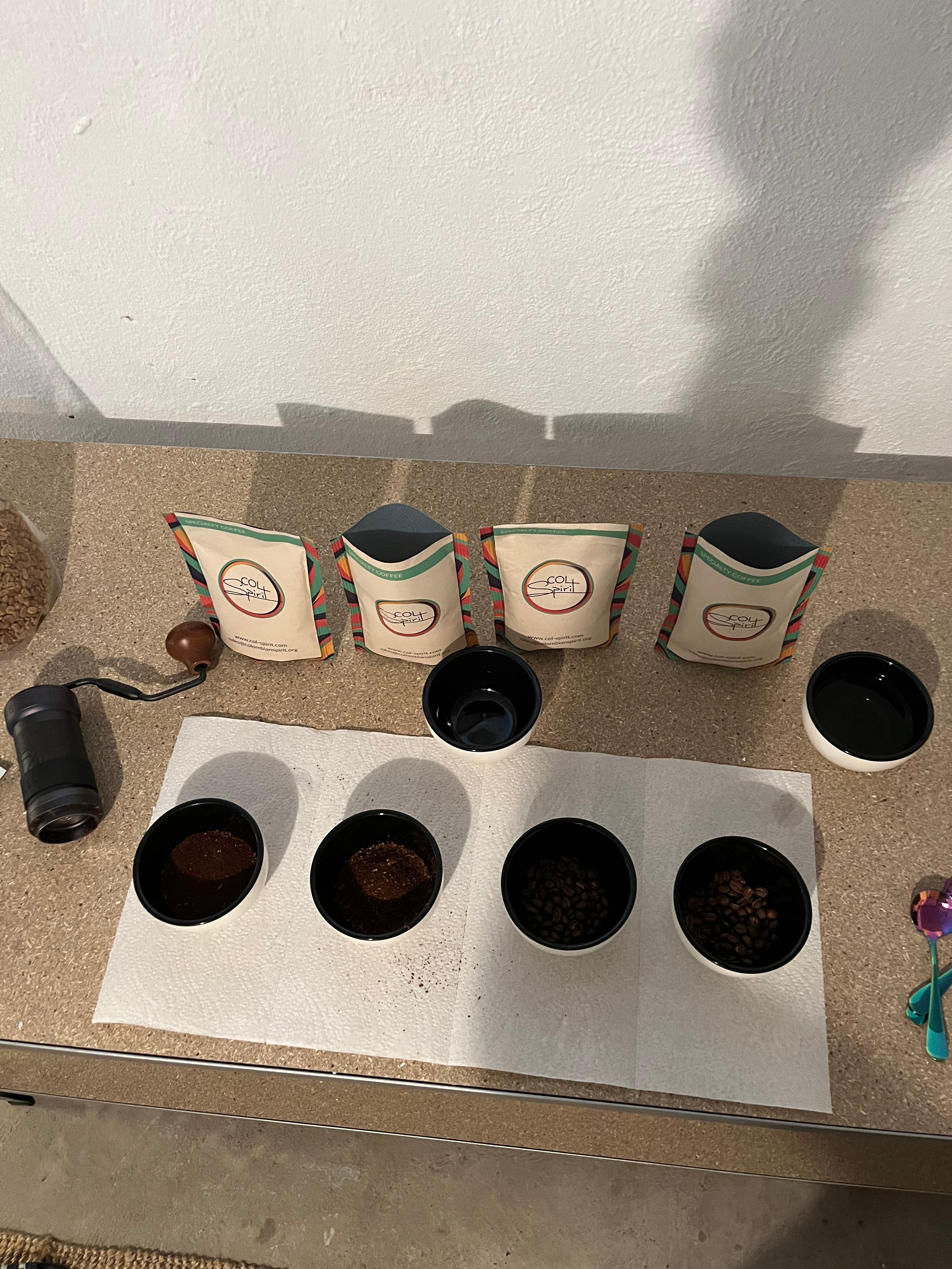 Cuppingbecher gefüllt mit gemahlenem Kaffee, davor Rohkaffeetüten (klein) links davon Handmühle.